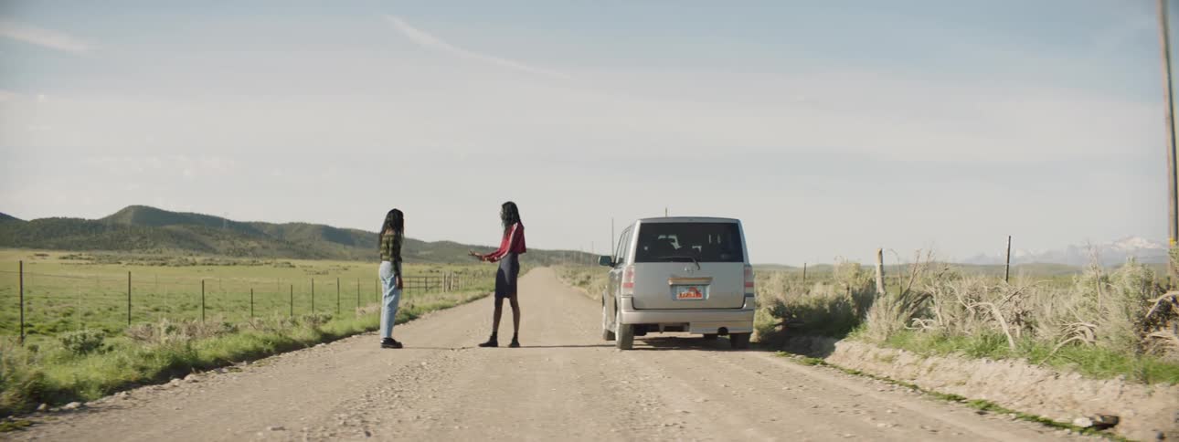 Two women argue on a country road next to a parked vehicle
