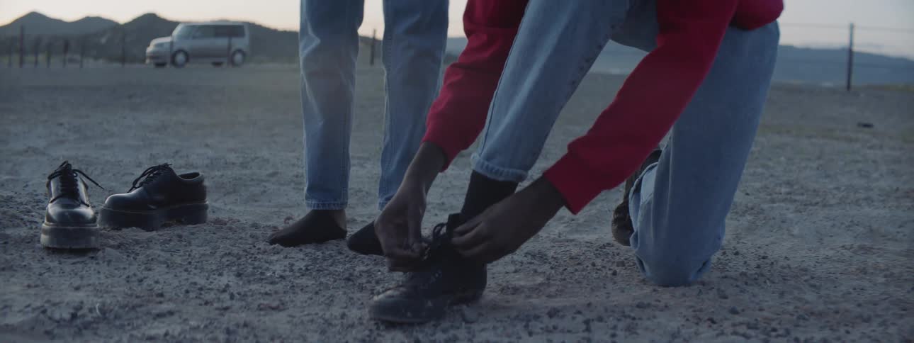 A person unties their shoe in the desert while a person wearing no shoes stands next to them