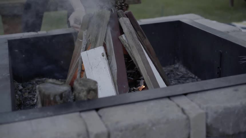 A woman in a hat sets fire to wood stacked into a tipi with a long match