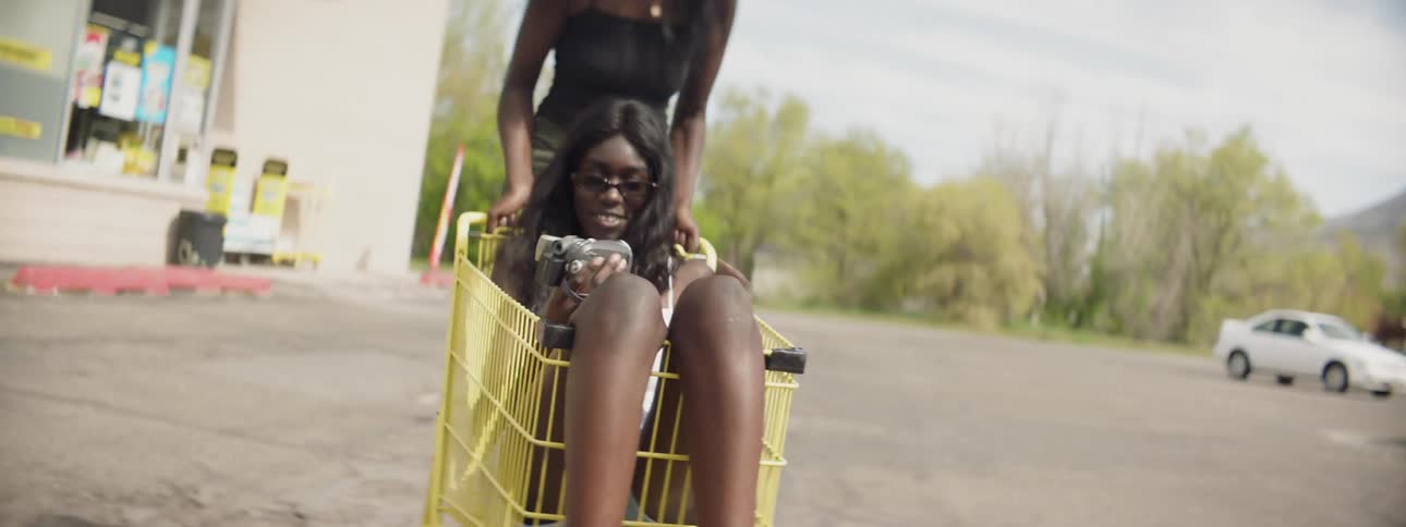 A woman pushes another woman in a shopping cart through a parking lot