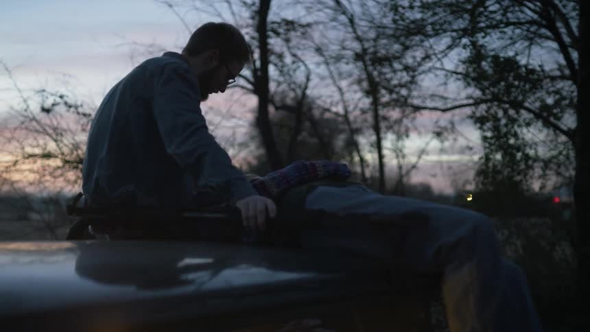 A man sits on top of a car with another person as cars pass on the road beside them