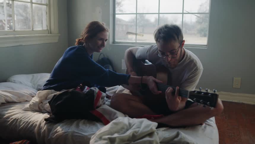 A man playing guitar for a woman sitting on his bed