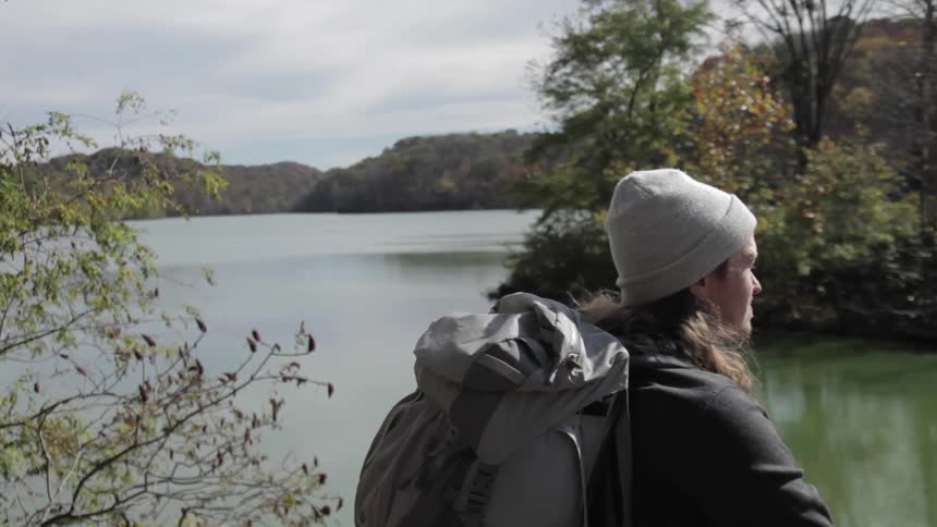 A young man, wearing a large backpack, sits next to a body of water