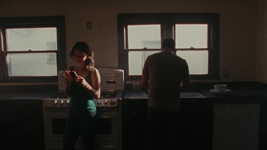 A woman leans against a kitchen stove and looks at her phone while a man works at the counter