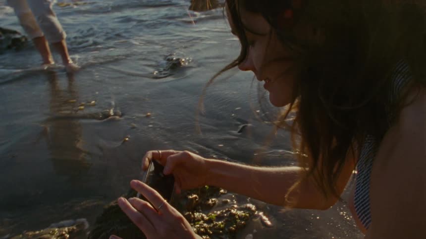 A young woman bending over to photograph something in the water as waves crash ashore and around rocks