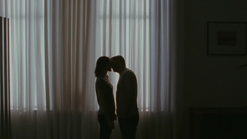 A man and woman share an intimate moment standing in front of a large curtained window