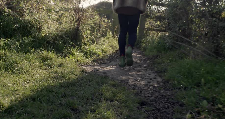 A young woman walking on a path through an area overgrown with weeds and headed towards an opening in a fence