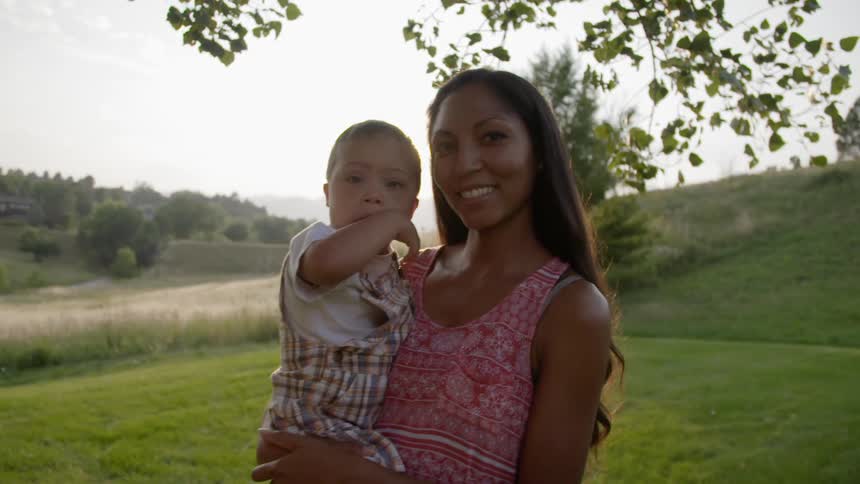 A woman standing on a green hilltop at sunset holds her toddler in her arms and smiles at the camera