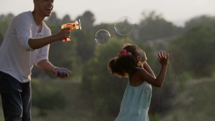 A father makes bubbles in the air while his daughter tries to catch them