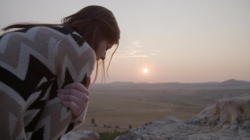 An unhappy woman stands high above a desert landscape and clutches her arms around herself