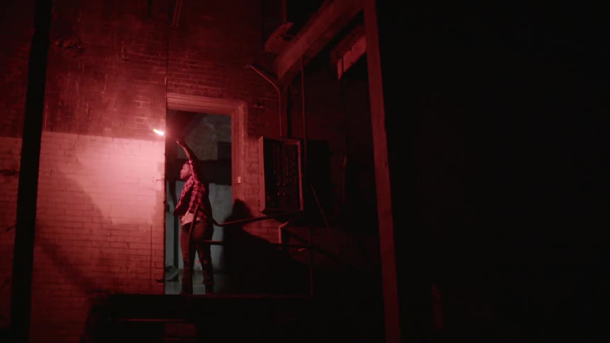 A woman uses a red flare to light up the doorway of a building at night so she can look at the wall