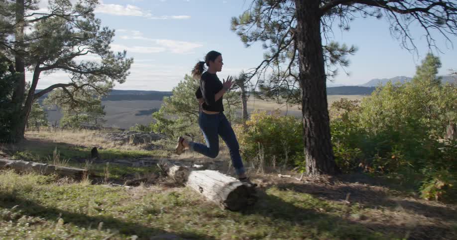 A woman jumps over a log as she runs through the woods