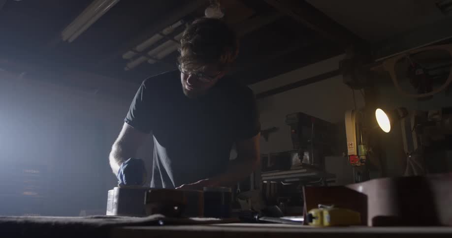 A man stains and polishes a wooden instrument in a garage