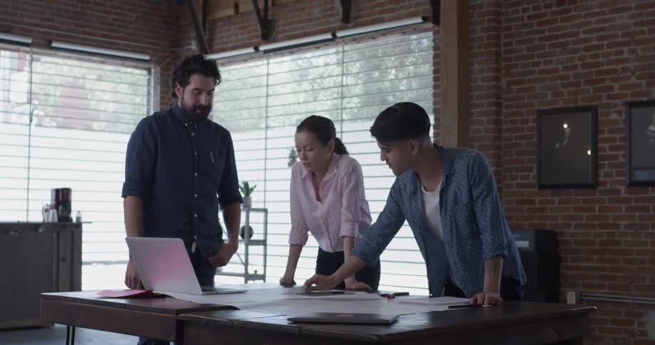 A woman and two men stand around a table with a computer on it and talk