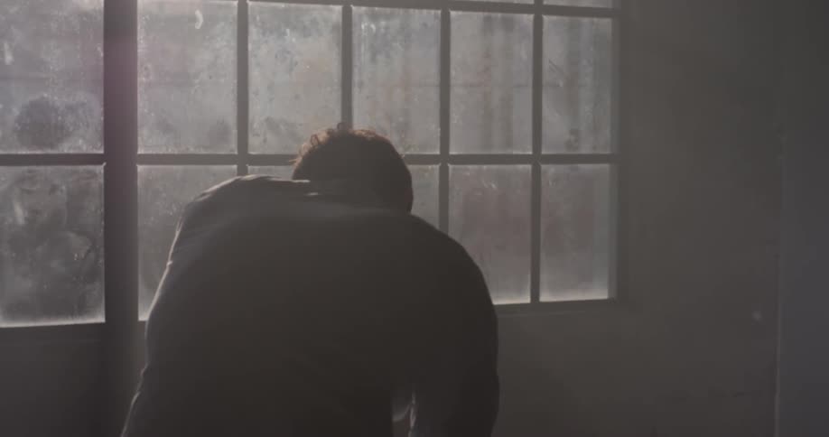 A man is slouched over in a dark room next to the window