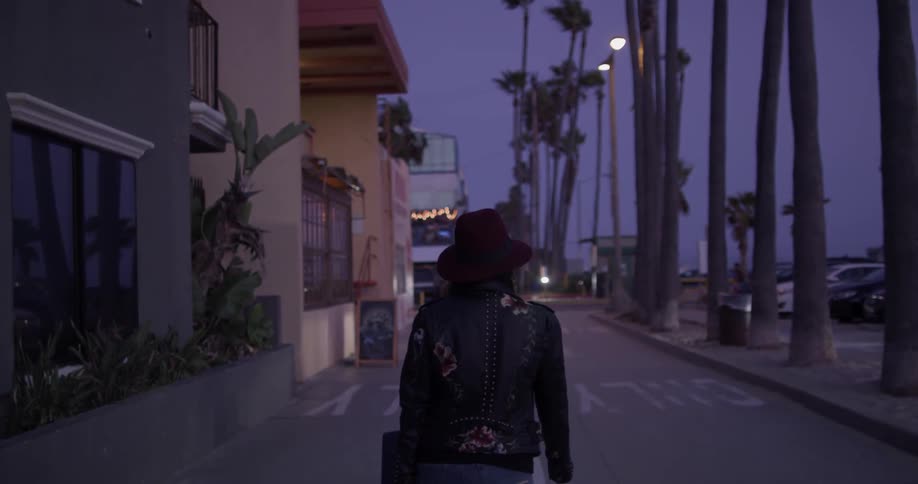 A woman carrying a guitar walks down a street at night