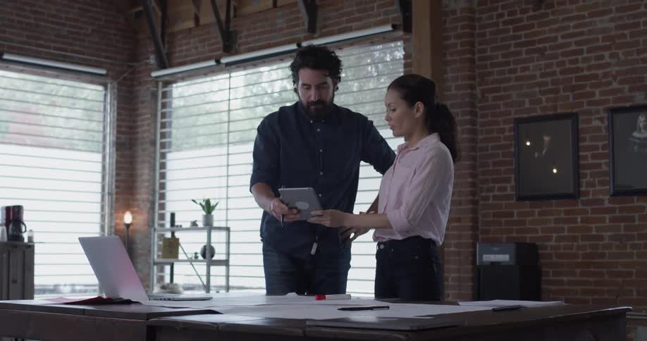 A woman shows a tablet to a man who is writing something on a paper and they look at it together