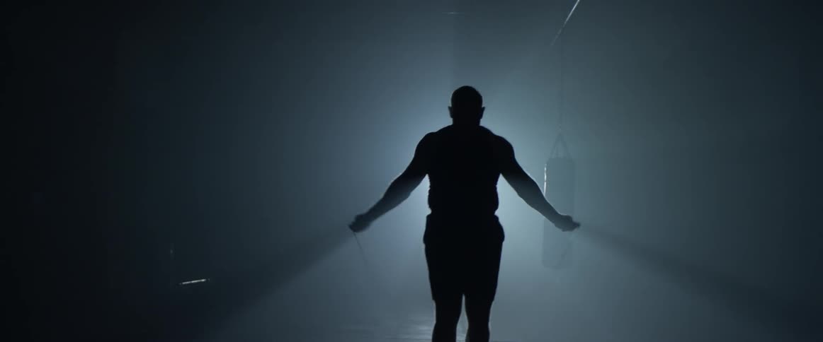 Man jump ropes inside a dim lit room with a spotlight behind him which he eclipses