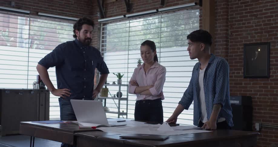 Two men and a woman discuss something over a table covered with papers and a laptop