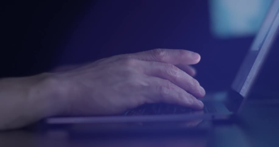 Man types on a laptop while sitting in a dark room