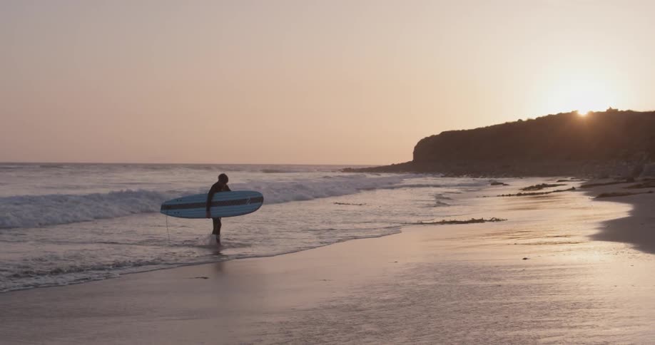 A man walks out of the ocean with a surfboard under his arm