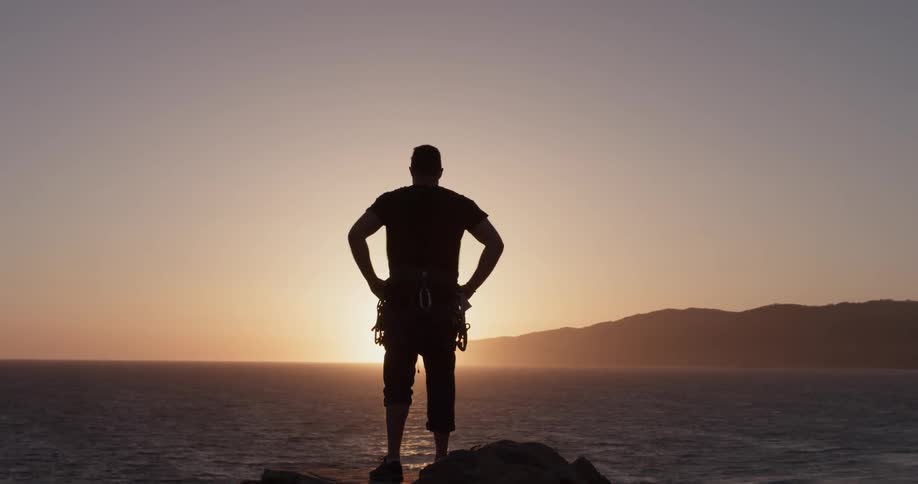 A man stands in front of the sun while on top of a rock overlooking a great body of water