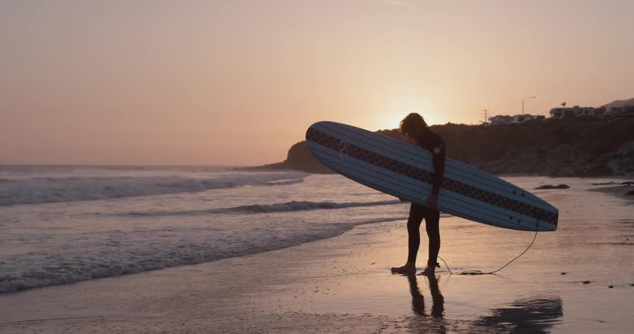A surfer stands with his board under his arm looking out at the ocean during a sunset