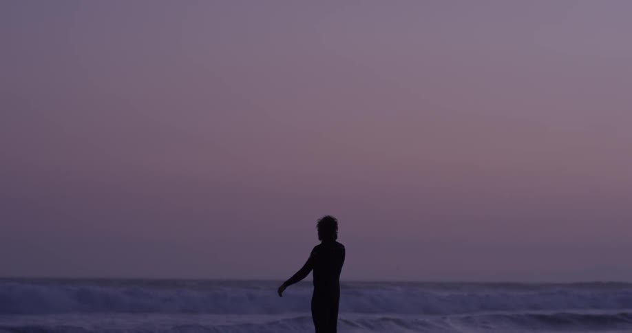A man walks on the beach wearing a wet suit as the sun sets or rises and waves come crashing in
