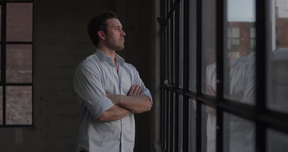 A man in a white shirt crosses his arms as he looks out a window
