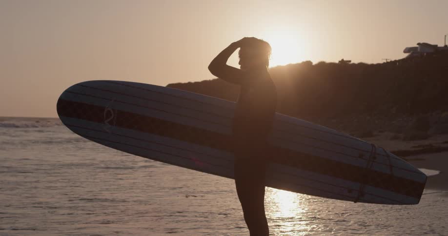 A surfer stands with his board under his arm during a sunset