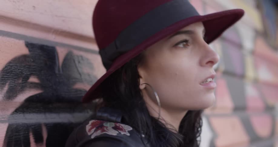 A woman wearing a hat with long black hair is singing and playing guitar up against a garage door with graffiti