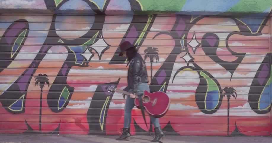 A woman walks by some graffiti on a garage door while holding a guitar and wearing a hat