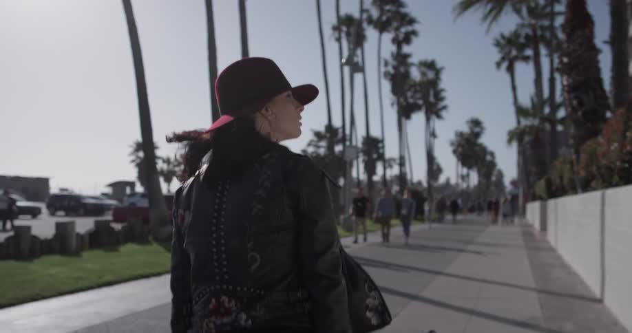 A woman wearing a hat and a leather jacket carries a guitar in a case down the street