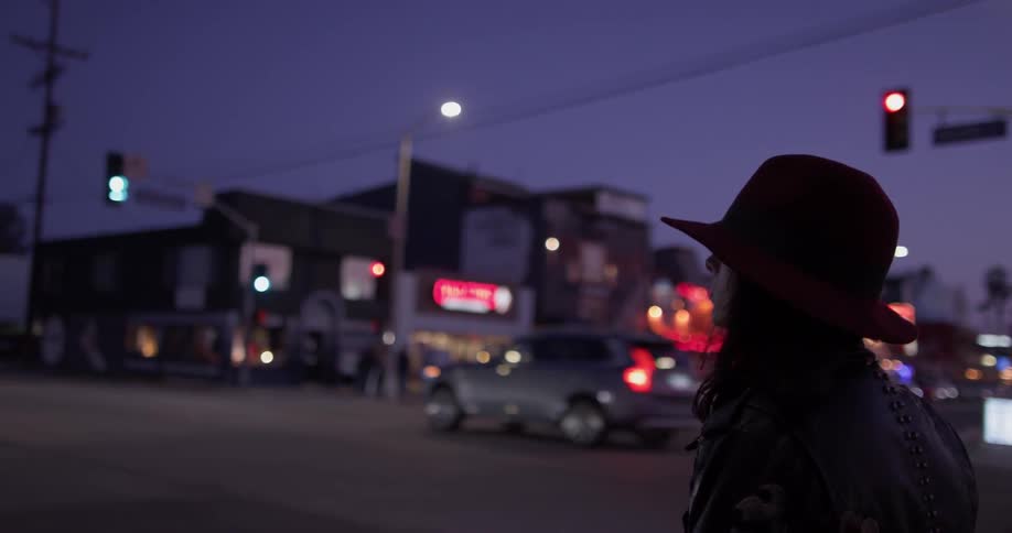 A woman wearing a hat stands on a street corner in the dark
