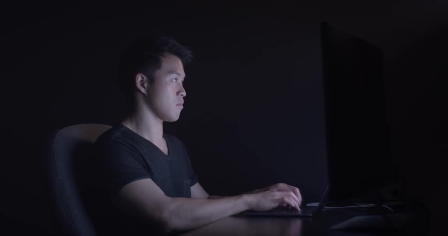 A man typing on a keyboard inside a dark room with the only light coming from the monitor