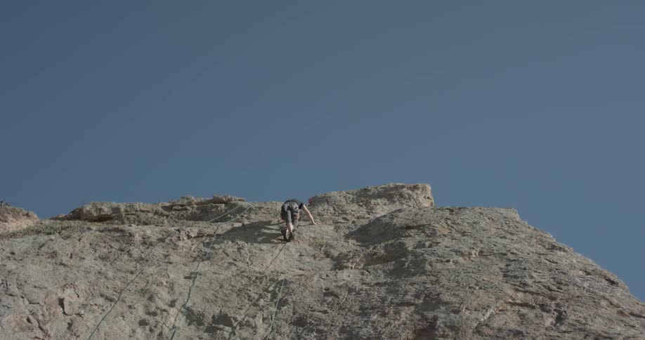 A man is rock climbing scaling a tall rock with the blue skies ahead of him