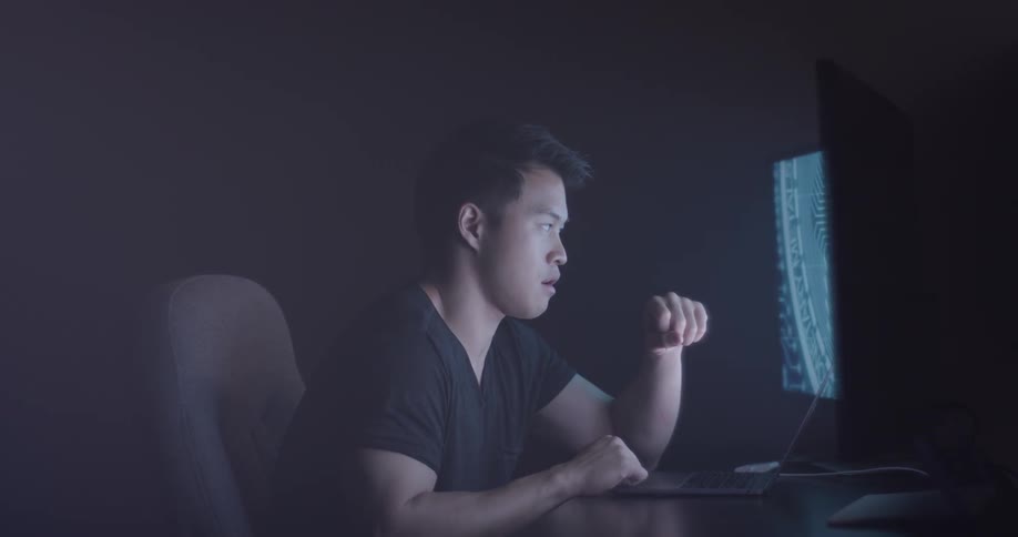 An asian man sits at his computer desk and his left monitor shows a bitcoin logo