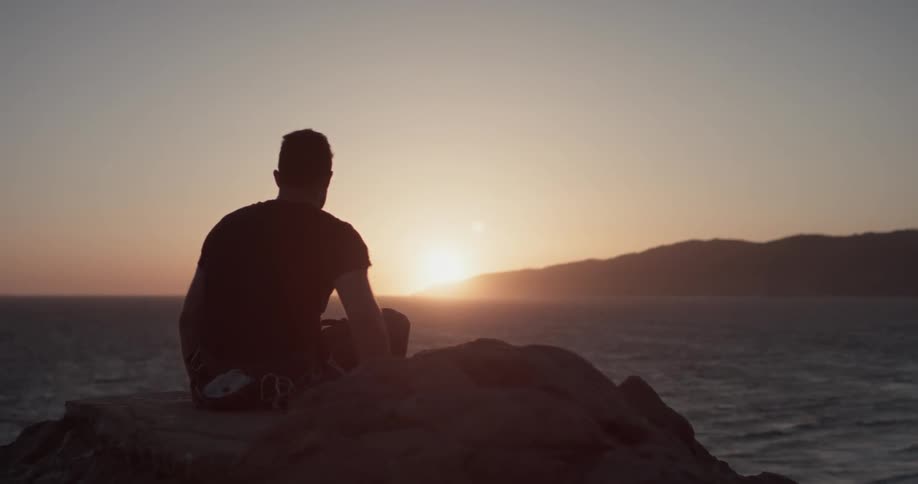 A man is sitting outside while the sun sets