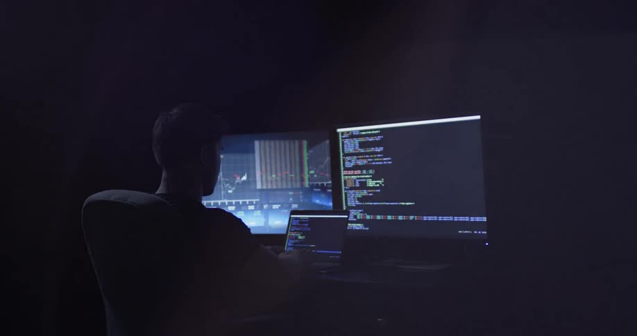 A man sits in the darkness illuminated by two computer monitors