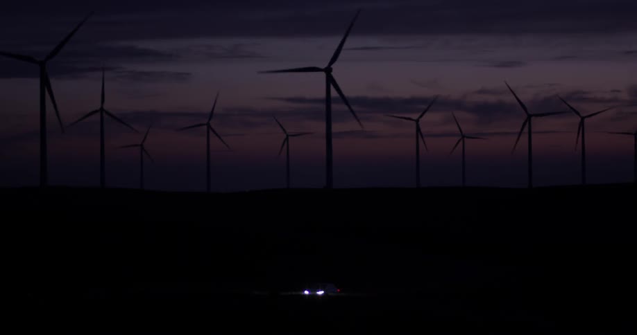 Windmills turn at night, with a car driving with headlights on in the foreground
