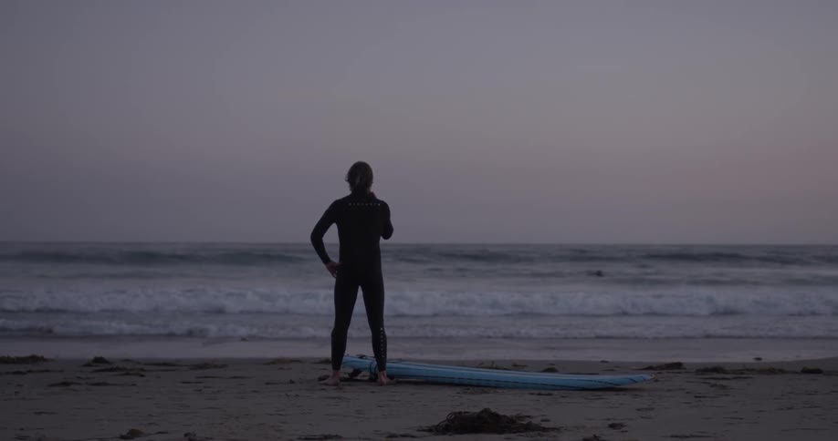 Surfer stands watching the ocean on the beach at sunset