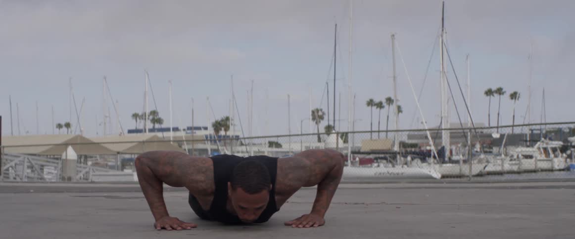 A large muscular man does push up tricks in front of a marina