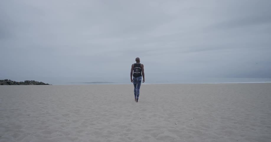 A man wearing leggings and a tank top walks on the beach towards the water on an overcast day