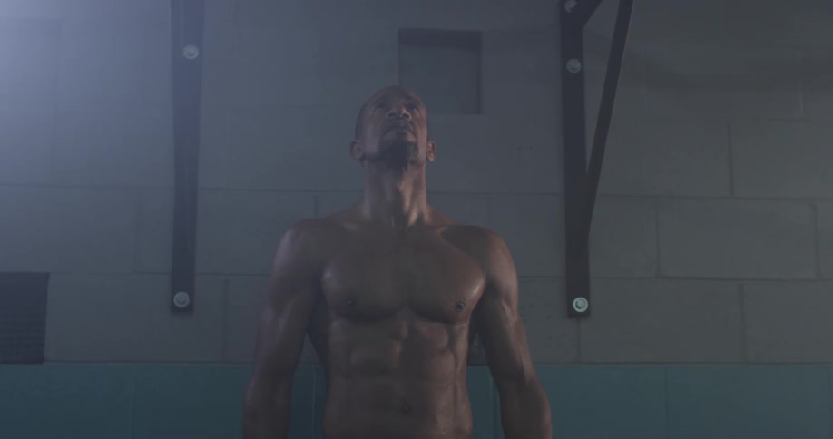 A very buff older man looks up at the pull up bar at the gym with a spotlight on him