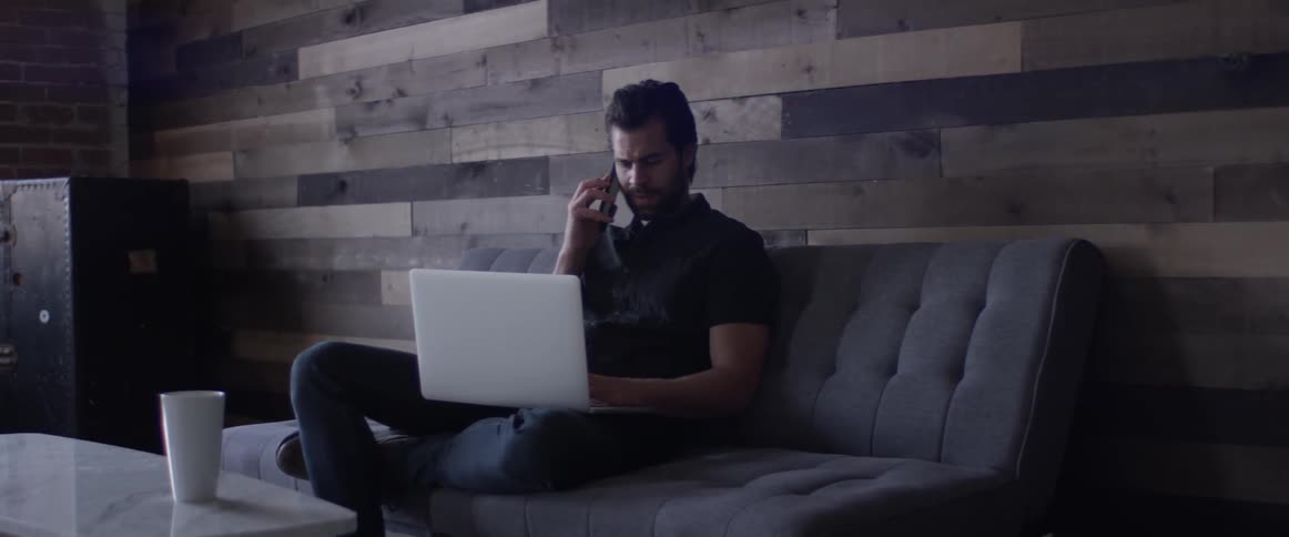 A man on a laptop reaches forward to pick up a cell phone and talk
