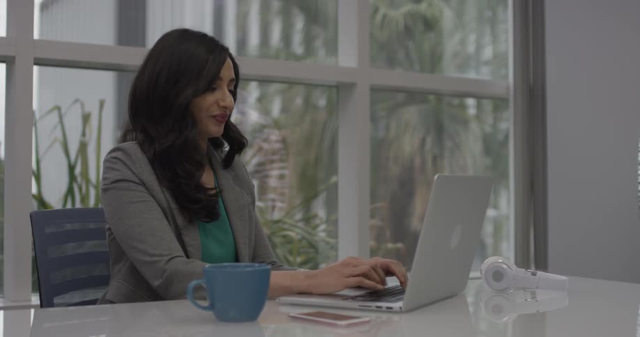 A woman sits at a conference table typing on her laptop with large paned windows behind her