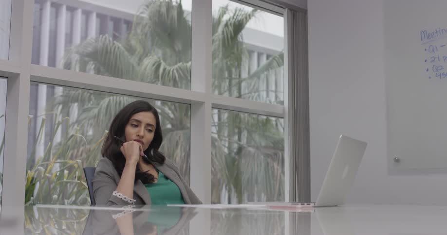A woman sits at a table in front of her laptop and thinks before writing on some paper