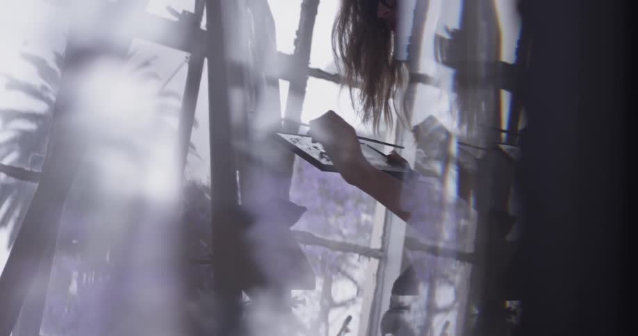 A woman wearing glasses stands at an easel and paints in front of a large window