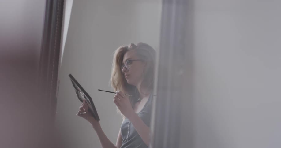 A beautiful nerdy black rim glasses wearing long haired women girl is painting with a paint brush inside