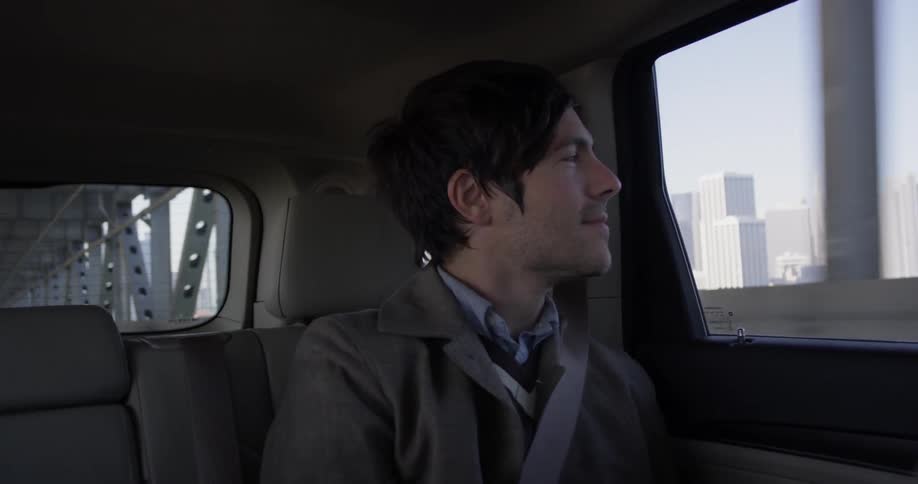 A man smiles and looks out a window, while riding in the backseat of a car